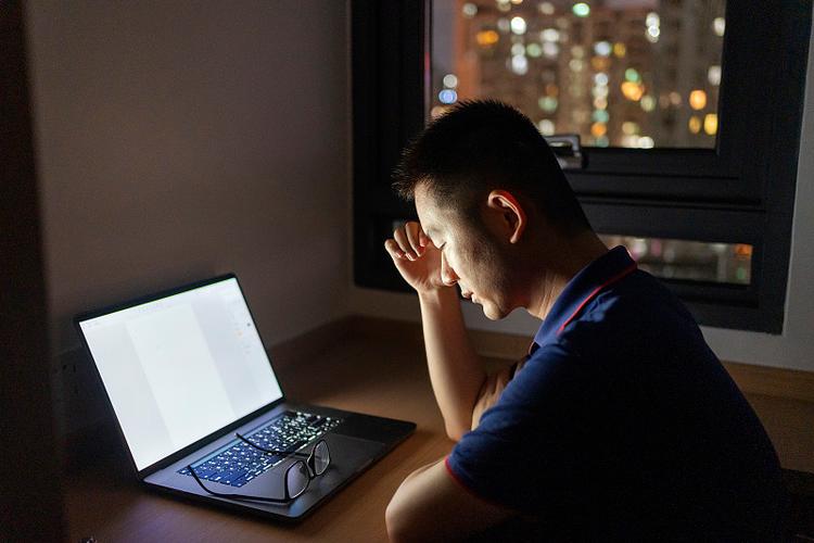 A tired man slumped at a laptop late at night, city lights in the window behind him